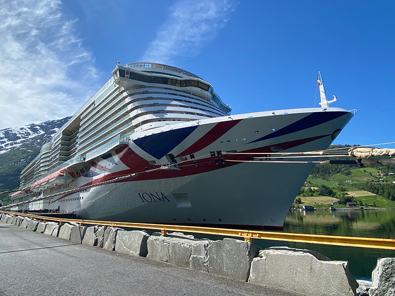 &O Cruises Iona docked beside a stone quayside in Olden. The enormous white cruise ship features a red and blue Union Jack design across its bow with green hills and snow capped mountains behind it under a bright blue sky.