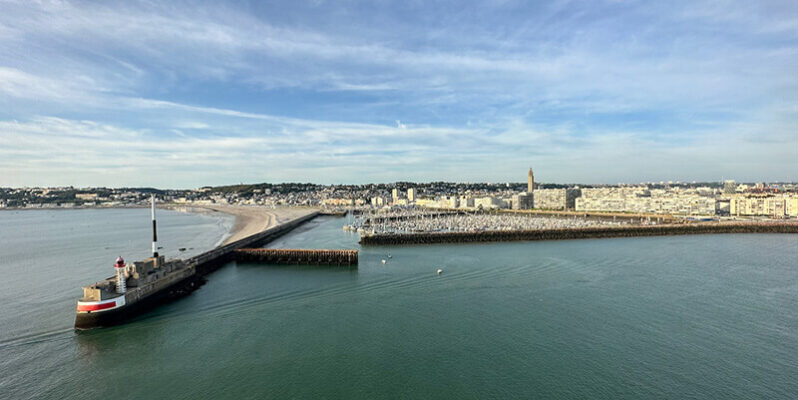 Elevated view over a Le Havre harbour with a long stone breakwater extending into calm blue green water and a small red and white lighthouse at its end. The town stretches along the coastline beneath a partly cloudy sky.