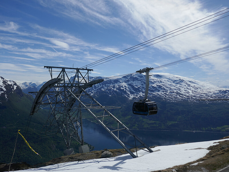 A black cable car hangs above a deep blue Norwegian fjord beside a large metal support tower. Snow covered mountains stretch across the background beneath a bright blue sky with wispy white clouds.