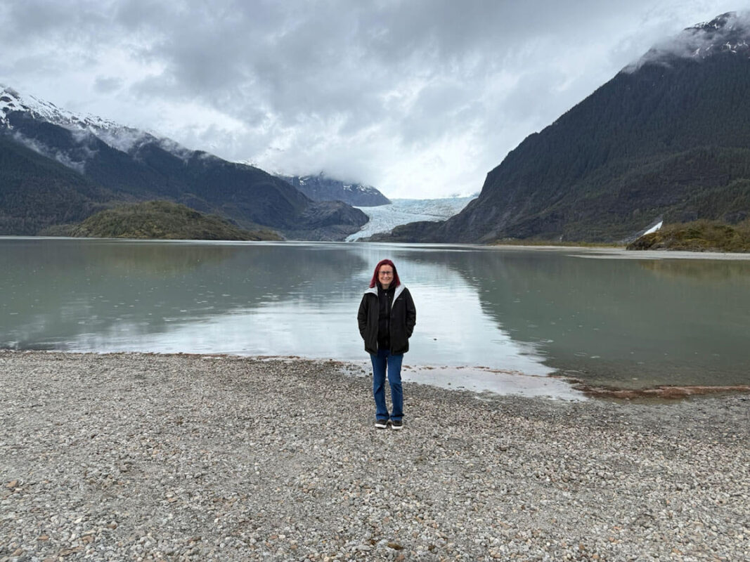 Emmaonline with red hair wearing a black jacket and blue jeans stands on a rocky shoreline in Alaska. Behind her is a calm glacial lake surrounded by towering mountains with a the Mendenhall Glacier visible in the distance.