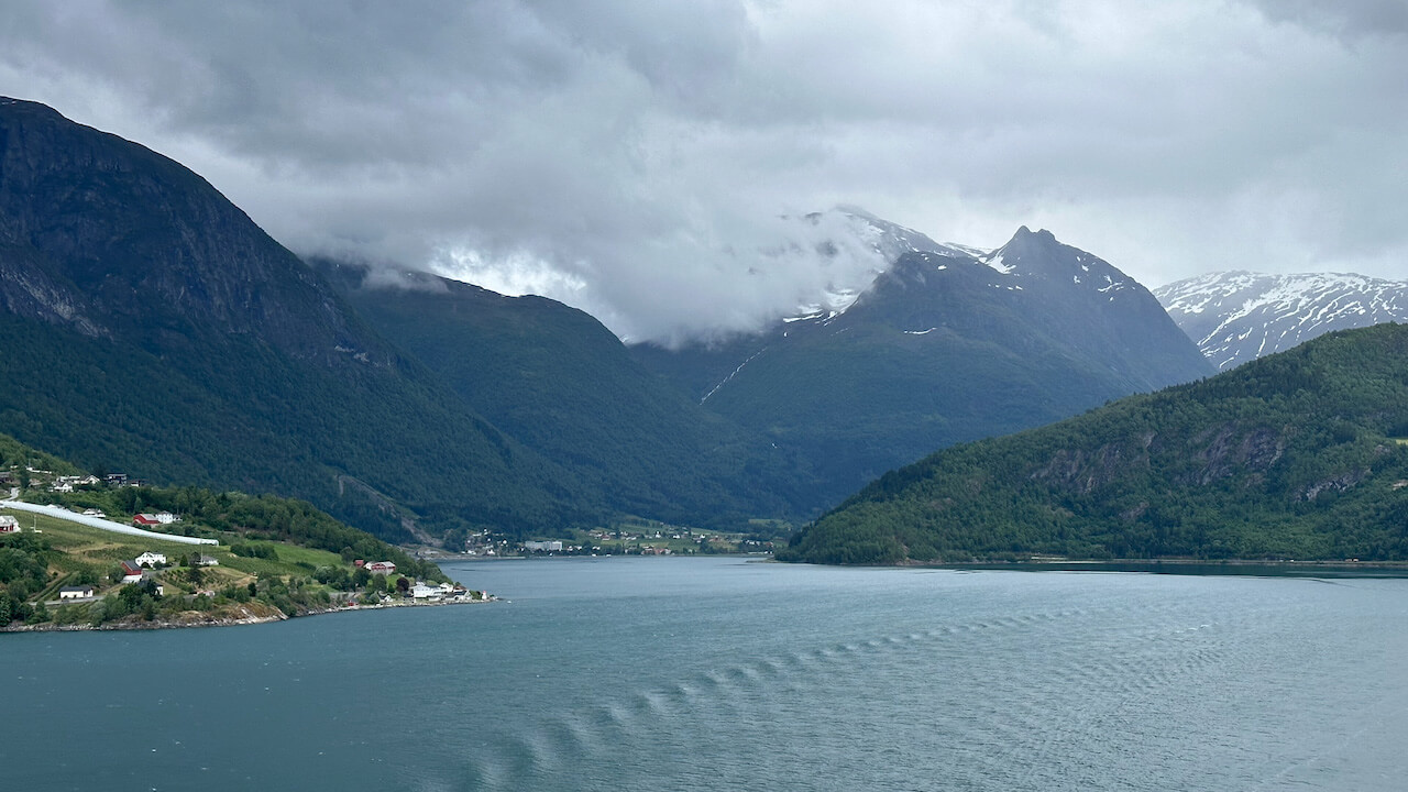 View across a Norwegian fjord towards towering green mountains with snow covered peaks partly hidden by low clouds. Small white and red houses dot the shoreline beside calm blue grey water beneath an overcast sky.