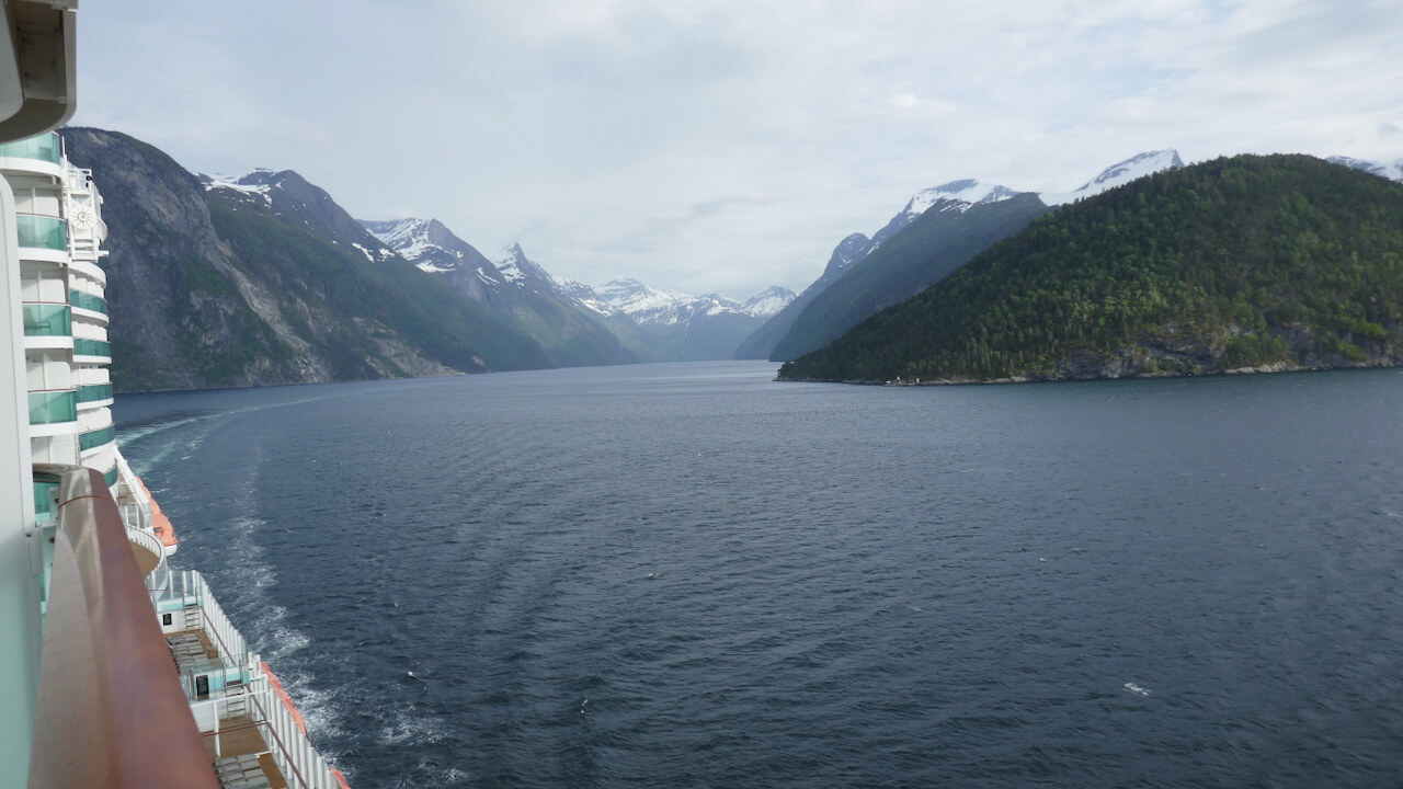 View from the balcony of a cruise ship sailing through a Norwegian fjord with deep blue water stretching between towering green mountains and snow covered peaks. The side of the ship and its wake are visible on the left as the fjord winds into the distance beneath a cloudy sky.