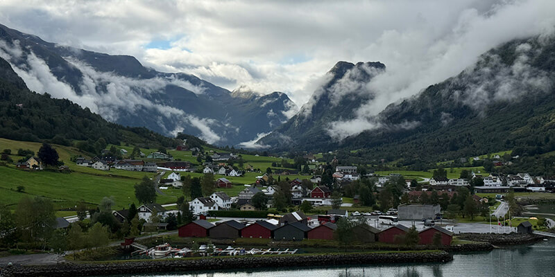 Olden, a small Norwegian village with colourful houses, green fields and red waterfront buildings sits in a valley surrounded by towering mountains. Thick white clouds drift between the mountain peaks creating a dramatic misty landscape.