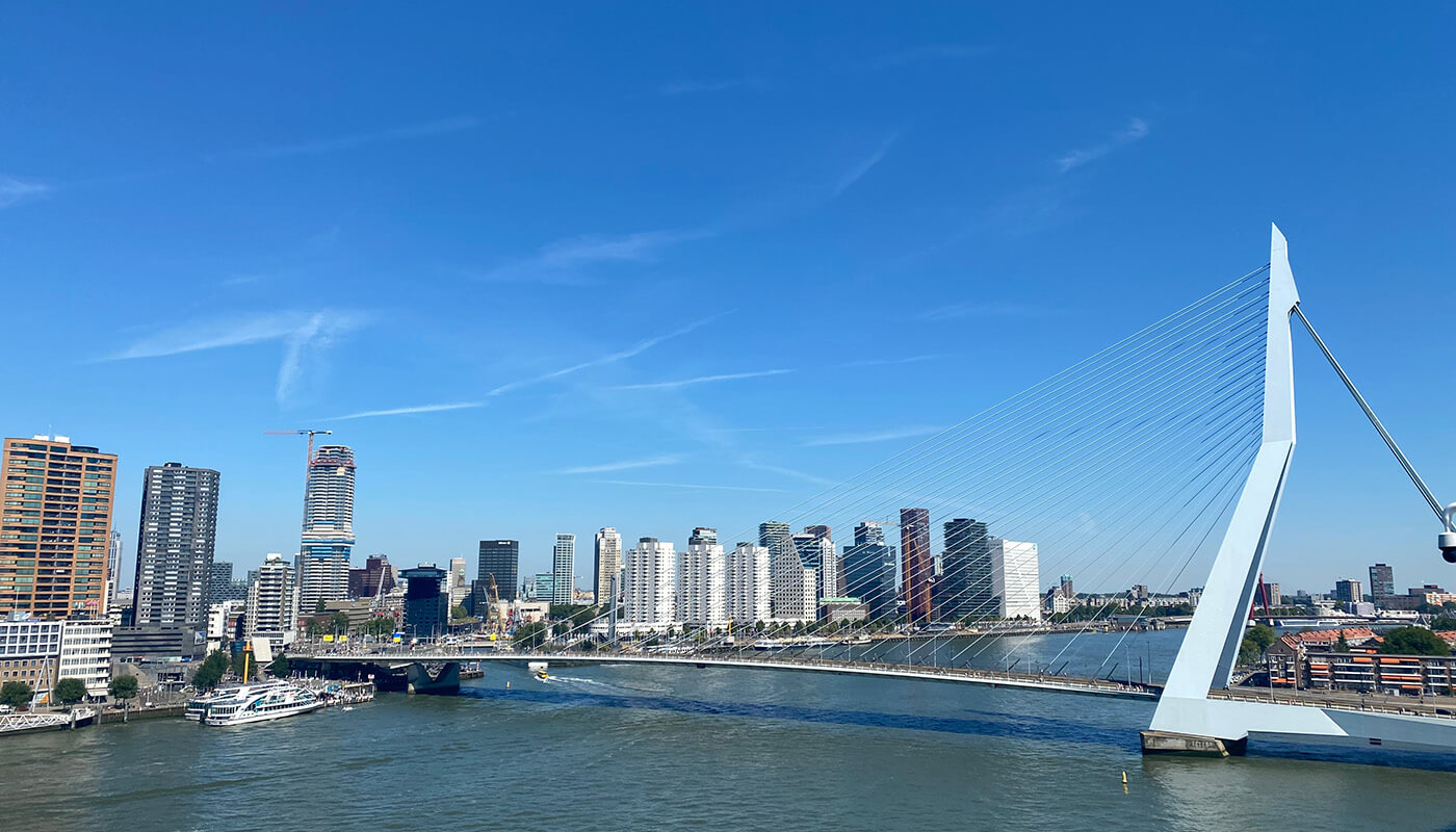 The white Erasmus Bridge stretches across the Nieuwe Maas river in Rotterdam with its tall angled pylon and cables creating a striking shape against the blue sky. Modern skyscrapers line the waterfront behind the bridge.