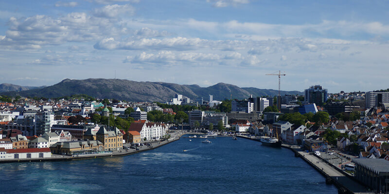 View across the harbour in Stavanger Norway with colourful waterfront buildings, boats and modern city buildings lining both sides of the water. Low rocky hills stretch across the horizon beneath a partly cloudy blue sky.