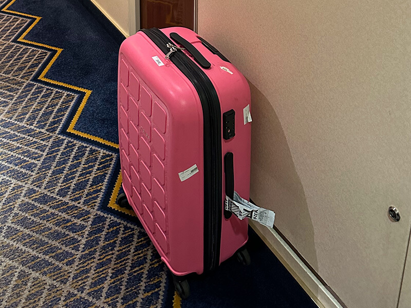 Bright pink hard shell suitcase standing upright in a cruise ship corridor beside a cabin door. The suitcase has four wheels and attached luggage tags and sits on a blue patterned carpet after being delivered to the cabin.