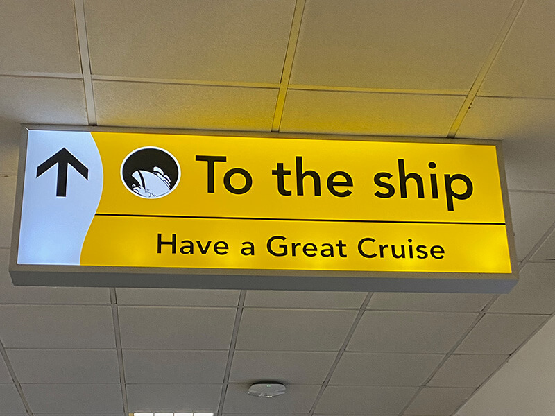A bright yellow overhead sign at a cruise terminal reads "To the ship" with an upward pointing arrow and cruise ship symbol. Below it are the words "Have a Great Cruise" welcoming passengers as they head towards their ship.