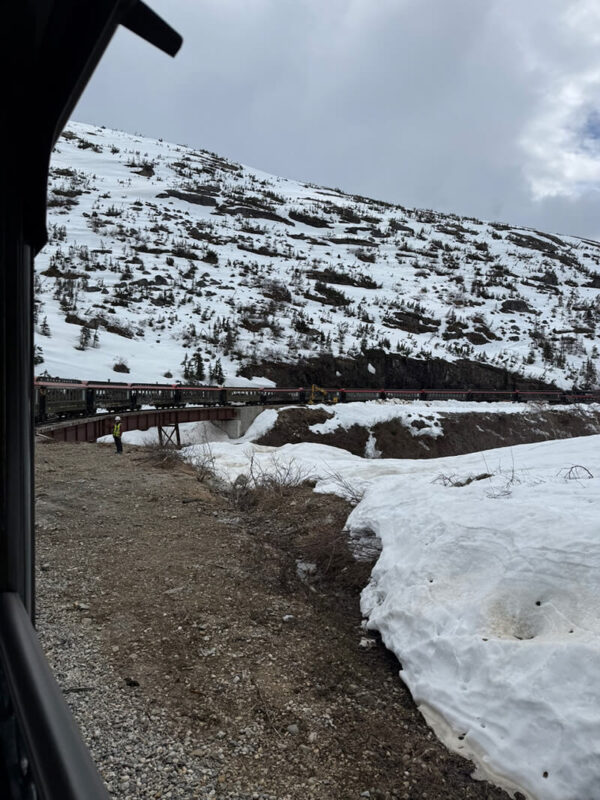 View from the back of the White Pass Railway as the train is crossing a bridge surrounded by snow covered hills and rocky terrain. The overcast skies and lingering snow show a colder side of an Alaska cruise excursion.